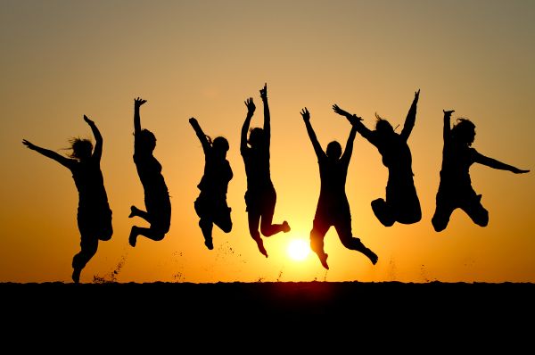 Group of seven people jumps as they are silhouetted against a sunset.